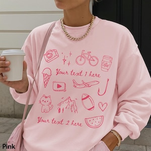 May include: Pink crewneck sweatshirt with various pink graphic illustrations, including a camera, bicycle, coffee cup, ice cream cone, and a cat. The sweatshirt has the text "Your text 1 here" and "Your text 2 here".