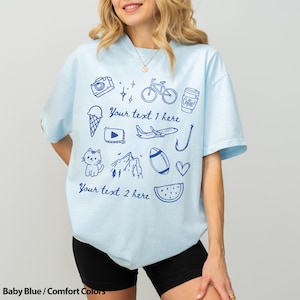 May include: Baby blue t-shirt with various blue doodles, including a camera, bicycle, coffee cup, ice cream cone, and a cat. The shirt has the text "Your text 1 here" and "Your text 2 here". The shirt is a comfort color.