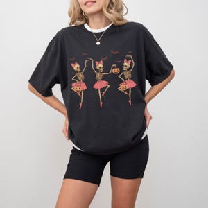 K&ouml;nnte beinhalten: Schwarzes T-Shirt mit einem Halloween-Motiv von drei tanzenden Skeletten in rosa Tutus, die K&uuml;rbislaternen halten. Das Design umfasst rosa Schleifen und Flederm&auml;use. Das T-Shirt hat einen wei&szlig;en Rundhalsausschnitt.