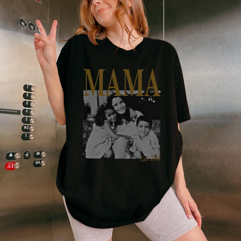 Puede incluir: Camiseta negra con la palabra "MAMA" en letras doradas sobre una foto en blanco y negro de una madre y dos hijos. La foto es rectangular y tiene el texto "Est. 2015" en la parte inferior.