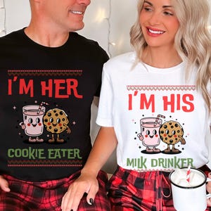 May include: Black and white t-shirts with matching graphics. The black shirt says "I'M HER COOKIE EATER" with a cartoon glass of pink milk and a cookie. The white shirt says "I'M HIS MILK DRINKER" with a cartoon glass of pink milk and a cookie.