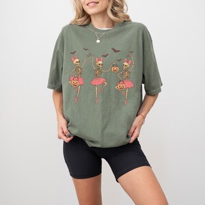 K&ouml;nnte beinhalten: Olivgr&uuml;nes T-Shirt mit einer Grafik von drei Skelett-Ballerinas in rosa Tutus, die K&uuml;rbis-Eimer halten und rosa Schleifen tragen. Flederm&auml;use sind &uuml;ber den Ballerinas. Das Shirt wird mit schwarzen Shorts getragen.