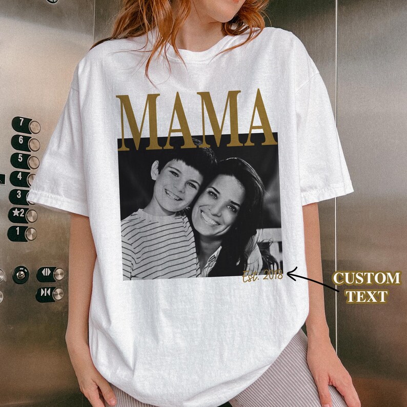 Puede incluir: Camiseta blanca con la palabra "MAMA" en letras doradas sobre una foto en blanco y negro de una madre y su hijo. La foto est&aacute; enmarcada por un rect&aacute;ngulo negro. El texto "Est. 2018" est&aacute; debajo.