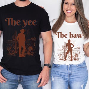 Op de afbeelding: Zwarte en witte T-shirts met een bruine afbeelding van een cowboy in een woestijnlandschap. Het zwarte shirt heeft "The yee" en het witte shirt "The haw" in bruine tekst. De shirts worden door twee personen gedragen.