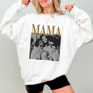 Puede incluir: Sudadera blanca con la palabra "MAMA" en letras doradas sobre una foto en blanco y negro de una madre y dos hijos. La sudadera tiene cuello redondo y mangas largas.