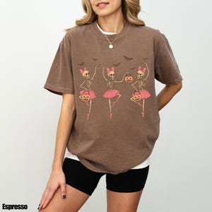 K&ouml;nnte beinhalten: Espressofarbenes T-Shirt mit einem Halloween-Design. Es zeigt drei Skelett-Ballerinas in rosa Tutus, die K&uuml;rbislaternen halten, mit Flederm&auml;usen dar&uuml;ber. Das T-Shirt hat eine lockere Passform.