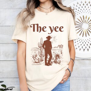 Op de afbeelding: Cr&egrave;mekleurig T-shirt met de tekst "The yee" in bruin. Het shirt heeft een bruine illustratie van een cowboy in een woestijnlandschap met cactussen en een ondergaande zon.