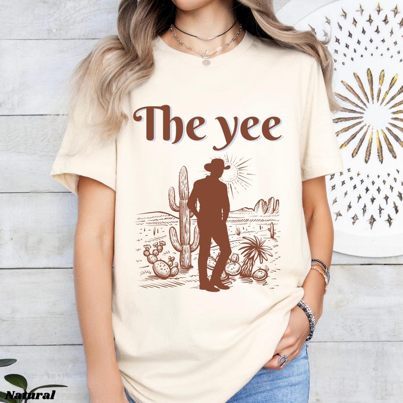 Op de afbeelding: Cr&egrave;mekleurig T-shirt met de tekst "The yee" in bruin. Het shirt heeft een bruine illustratie van een cowboy in een woestijnlandschap met cactussen en een ondergaande zon.