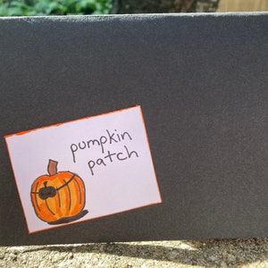May include: A white card with an orange and black hand-drawn pumpkin with an eye patch and the words "pumpkin patch" written in black marker.