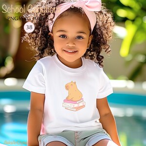 May include: A young child wearing a white t-shirt with a cartoon capybara sitting on books. The shirt has a pink and orange color scheme. The child is also wearing a pink headband.