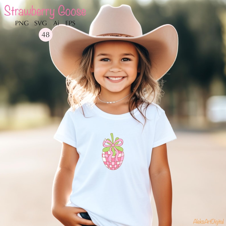 May include: A young girl wearing a white t-shirt with a pink and green strawberry design. She is wearing a brown cowboy hat and smiling.