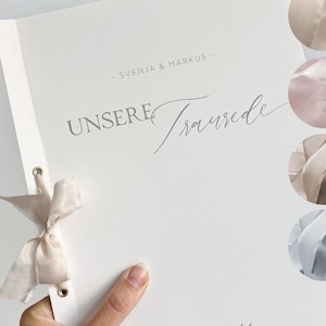 Könnte beinhalten: Ein weißes Programm für eine Hochzeitszeremonie mit dem Text "UNSERE Trauzed" und dem Datum "10. AUGUST 2025". Das Programm ist mit einer kleinen Zeichnung eines Zweigs mit Blättern verziert. Eine Hand hält das Programm, das mit einem hellrosa Band gebunden ist.