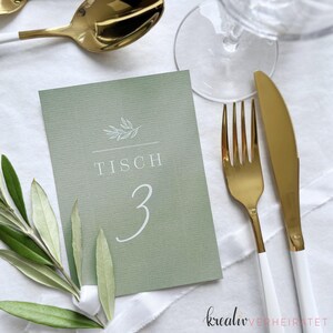Tischnummer als Karte aus Stukturpapier in der der Farbe Eukalyptus/Salbei. Die Karte mit weißem Aufdruck der Tischnummer und einer Olivenzweig-Grafik liegt auf einem weiß eingedecktem Tisch zur Hochzeit. Danebenliegt goldenes Besteck.