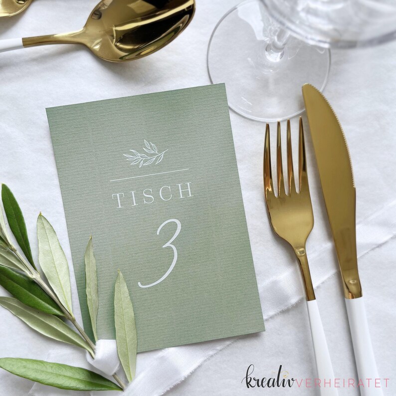 Tischnummer als Karte aus Stukturpapier in der der Farbe Eukalyptus/Salbei. Die Karte mit weißem Aufdruck der Tischnummer und einer Olivenzweig-Grafik liegt auf einem weiß eingedecktem Tisch zur Hochzeit. Danebenliegt goldenes Besteck.