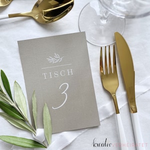 Die Karte mit gedruckter Tischnummer für Feiern aus farbigem Papier mit weißem Druck liegt auf einer weißen Tischdecke. Zu sehen ist goldenes Besteck sowie ein Glas. Die Schilder sind perfekt für Hochzeiten und als Tischdeko für die Hochzeitsfeier.