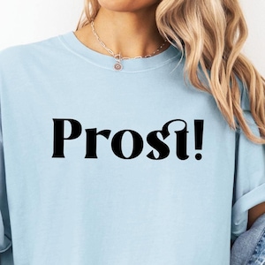 May include: A light blue t-shirt with the word "Prost!" printed in black on the front. The shirt is being worn by a person with long blonde hair.
