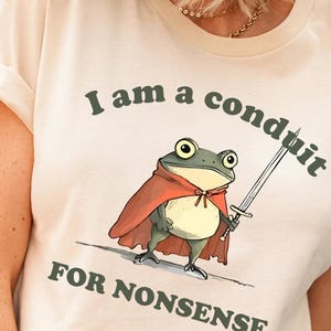 May include: Cream-colored t-shirt featuring a cartoon frog in a red cape holding a sword. The text "I am a conduit for nonsense" is printed above and below the frog illustration in a dark green color.