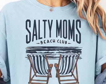 May include: Light blue t-shirt with the text "SALTY MOMS - BEACH CLUB-". The shirt features a graphic of two beach chairs and the phrase "Girls Just Wanna Have Sun".