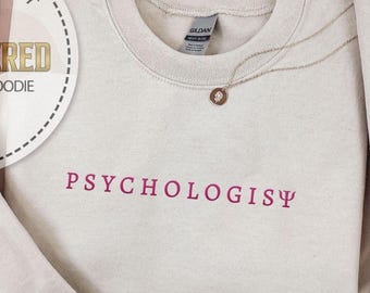 May include: A beige sweatshirt with the word "PSYCHOLOGIST" embroidered in a deep pink color. A gold necklace with a small pendant rests on the sweatshirt. The image also includes a circular logo that says "EMBROIDERED SHIRT & HOODIE".