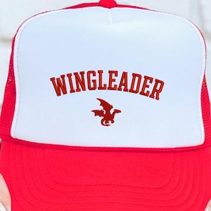 May include: Red and white trucker hat with the word "WINGLEADER" in red block letters and a red dragon graphic. The hat has a white front panel and a red brim and mesh back.