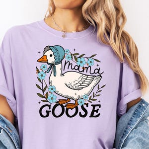 Puede incluir: Una camiseta de color lavanda con un gr&aacute;fico de una oca con un gorro azul y rodeada de flores. El texto "Mama Goose" est&aacute; impreso debajo de la oca.
