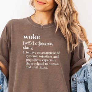 May include: A brown t-shirt with the word "woke" defined in white text. The definition reads: "[wōk] adjective, slang 1. to have an awareness of systemic injustices and prejudices, especially those related to human and civil rights."