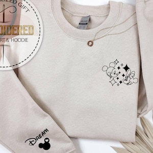 May include: Beige sweatshirt with a black Mickey and Minnie Mouse graphic and the word "DREAM" on the sleeve. A gold necklace with a pendant is draped on the sweatshirt. The sweatshirt is a Gildan brand.