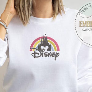 May include: White sweatshirt with a rainbow and the Disney castle with the word "Disney" underneath. The sweatshirt is being worn by a person.