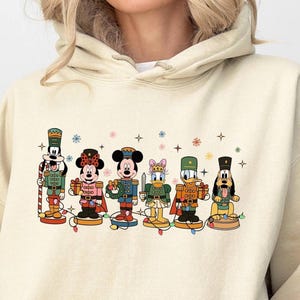 May include: A beige hoodie featuring a colorful graphic of Disney characters as nutcrackers. The characters include Mickey Mouse, Minnie Mouse, Donald Duck, and Goofy, each in festive outfits. Decorative snowflakes and lights are also present.