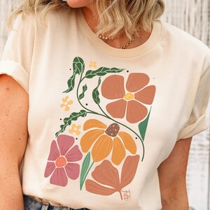 May include: A cream colored t-shirt with a floral design featuring orange, red, and pink flowers. The flowers are stylized and have a retro feel.