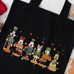 May include: Black tote bag featuring a colorful illustration of Disney characters dressed as toy soldiers. The characters include Mickey Mouse, Minnie Mouse, Donald Duck, and Pluto, with snowflake accents. The bag has black handles.