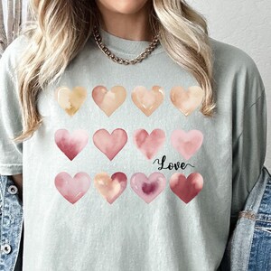 Puede incluir: Una camiseta verde claro con un dise&ntilde;o de acuarela de corazones rosas y melocot&oacute;n. La camiseta tiene la palabra "Love" escrita en letra negra en la parte delantera.