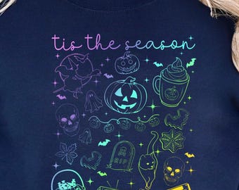May include: Navy blue sweatshirt with a colorful Halloween-themed graphic. The design includes pumpkins, bats, ghosts, skulls, and the phrase "tis the season" in a gradient font. A festive and spooky design.