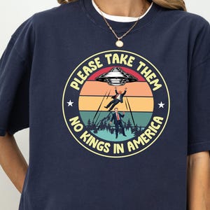 Op de afbeelding: Marineblauwe t-shirt met een cirkelvormige afbeelding. De afbeelding toont een UFO die een persoon ontvoert, met de tekst "PLEASE TAKE THEM, NO KINGS IN AMERICA". Het ontwerp heeft een retro zonsondergang kleurenschema.