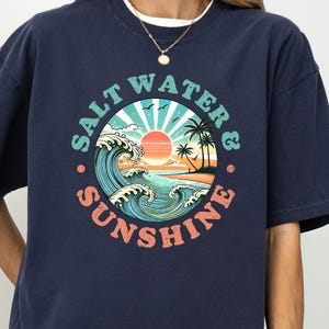 May include: Dark blue t-shirt with a circular graphic depicting a beach scene. The design includes the text "SALT WATER & SUNSHINE", waves, a sun, and palm trees. The shirt has short sleeves and a loose fit.