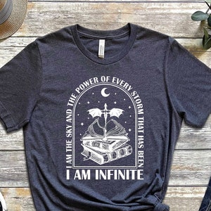 May include: A dark gray t-shirt with a white graphic design. The graphic features a crescent moon, a sword, a dragon, and a stack of books. The text on the shirt reads "I am the sky and the power of every storm that has been I am infinite."