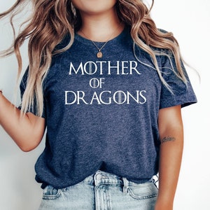 Op de afbeelding: Een marineblauw T-shirt met de tekst "MOTHER OF DRAGONS" in wit. Het shirt wordt gedragen door een persoon met lang blond haar en een blauwe jeans.