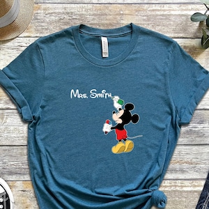 May include: A teal blue t-shirt with a white print of Mickey Mouse holding a paintbrush and a green paint can. The text "Mrs. Smith" is printed above Mickey Mouse.