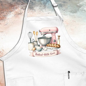 May include: White kitchen apron with a pink stand mixer, a loaf of bread, and other baking supplies. The apron has a pocket and the text "Baked With Love" on a pink ribbon.