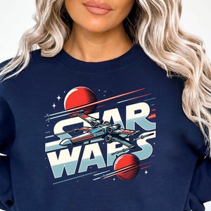 Puede incluir: Una sudadera azul marino con un gr&aacute;fico de una nave espacial volando por el espacio con el texto "Star Wars" en letras blancas y rojas.