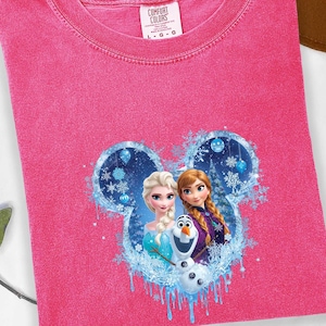 May include: A pink Comfort Colors t-shirt featuring a graphic of Elsa, Anna, and Olaf from Frozen, set within a Mickey Mouse-shaped design with snowflakes and blue accents. The shirt has a crew neck and a tag.