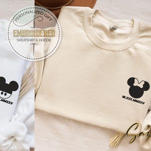 May include: White and beige sweatshirts with embroidered cartoon character designs. The white sweatshirt features a Mickey Mouse design, while the beige one has a Minnie Mouse design. Both have the text "BLXXX.MMXXV" below the designs.