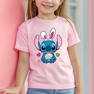 May include: A pink t-shirt with a blue cartoon character wearing bunny ears and holding an Easter egg. The character has a blue nose and big eyes. The shirt has a colorful heart design.