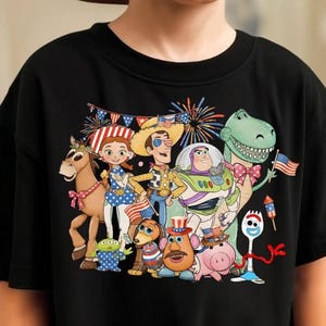 Puede incluir: Una camiseta negra de manga corta con un estampado gr&aacute;fico colorido de varios personajes de juguetes animados. Los personajes est&aacute;n decorados con patrones de banderas, estrellas y rayas, acompa&ntilde;ados de fuegos artificiales y banderines.