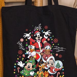 May include: Black tote bag featuring a festive Christmas design with Disney characters. Goofy, Daisy Duck, Donald Duck, Mickey Mouse, Pluto, and Minnie Mouse are dressed in holiday attire. The bag includes the characters' signatures and colorful Christmas lights.