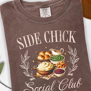 May include: Brown t-shirt with the text "SIDE CHICK Social Club" and an illustration of Thanksgiving side dishes. The design includes rolls, mashed potatoes, green beans, and cranberry sauce. The shirt is a Comfort Colors brand.