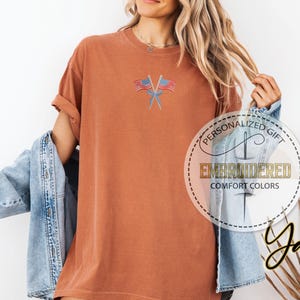 May include: Rust-colored t-shirt with embroidered American flags. The shirt has a relaxed fit and rolled-up sleeves. The text "PERSONALIZED GIFT EMBROIDERED COMFORT COLORS" is printed on a circular label.