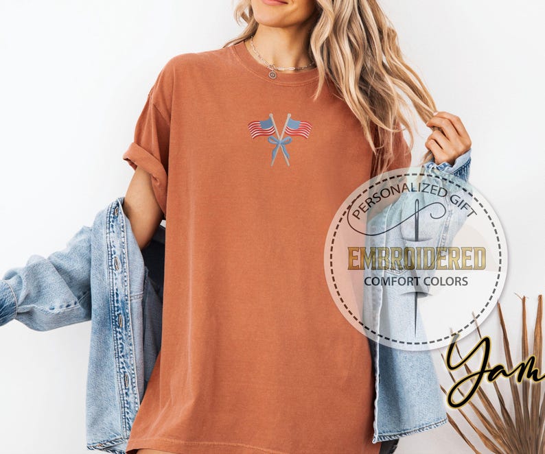 May include: Rust-colored t-shirt with embroidered American flags. The shirt has a relaxed fit and rolled-up sleeves. The text "PERSONALIZED GIFT EMBROIDERED COMFORT COLORS" is printed on a circular label.