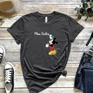 May include: A dark gray t-shirt with a white graphic of Mickey Mouse holding a pencil and a green apple. The text "Mrs. Smith" is printed above Mickey Mouse.