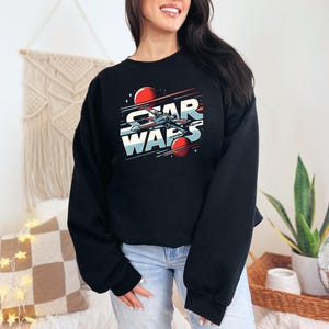 Puede incluir: Sudadera negra recortada con un gr&aacute;fico de una nave espacial volando por el espacio con el texto "Star Wars" en una fuente retro.